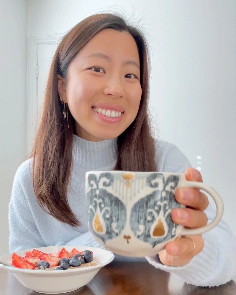 A person smiling, holding a patterned mug in one hand, while sitting at a table. In front of them is a bowl with blueberries and strawberries. They are wearing a light sweater against a plain background.